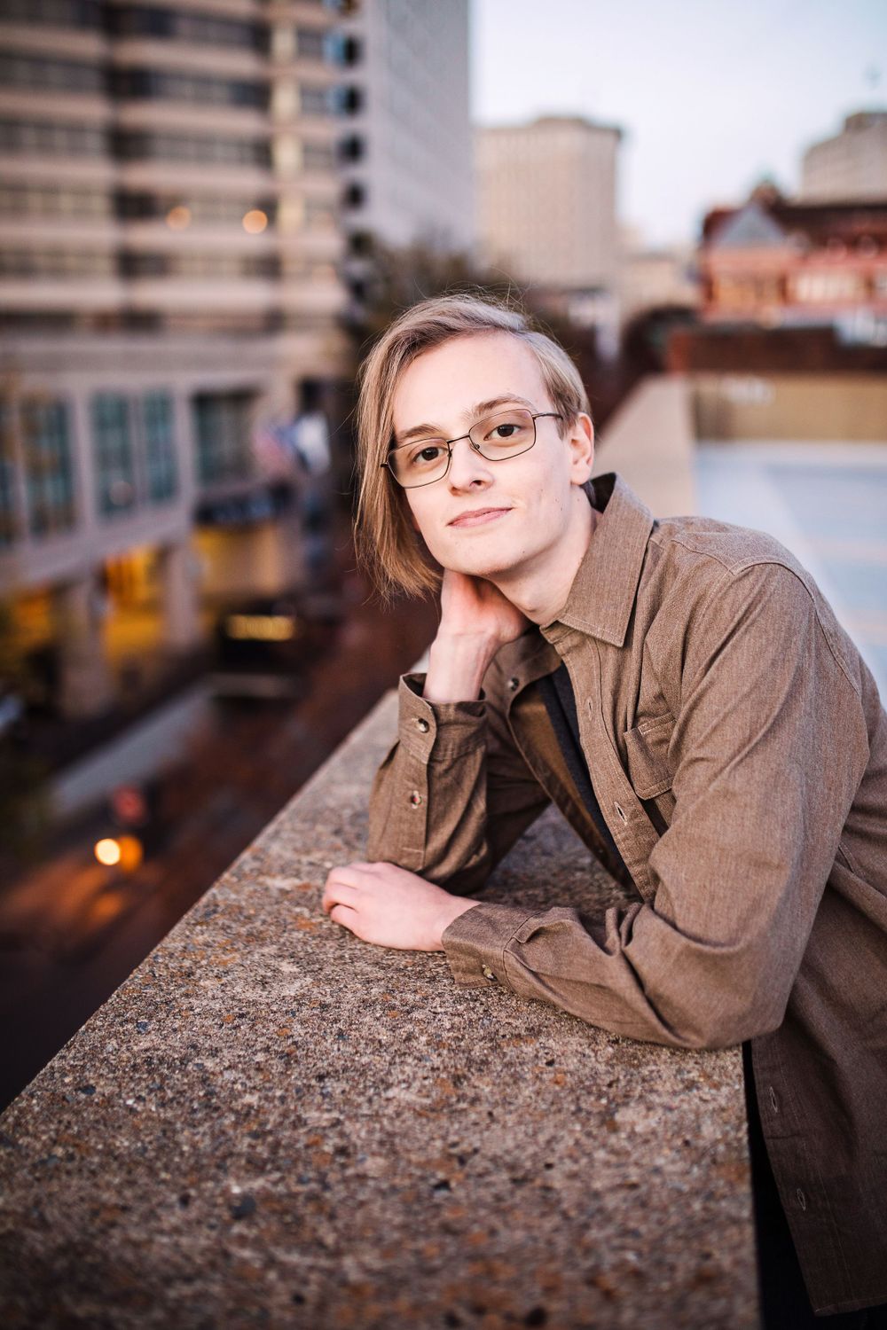Young person with glasses leaning on rooftop ledge with city skyline at dusk behind them.