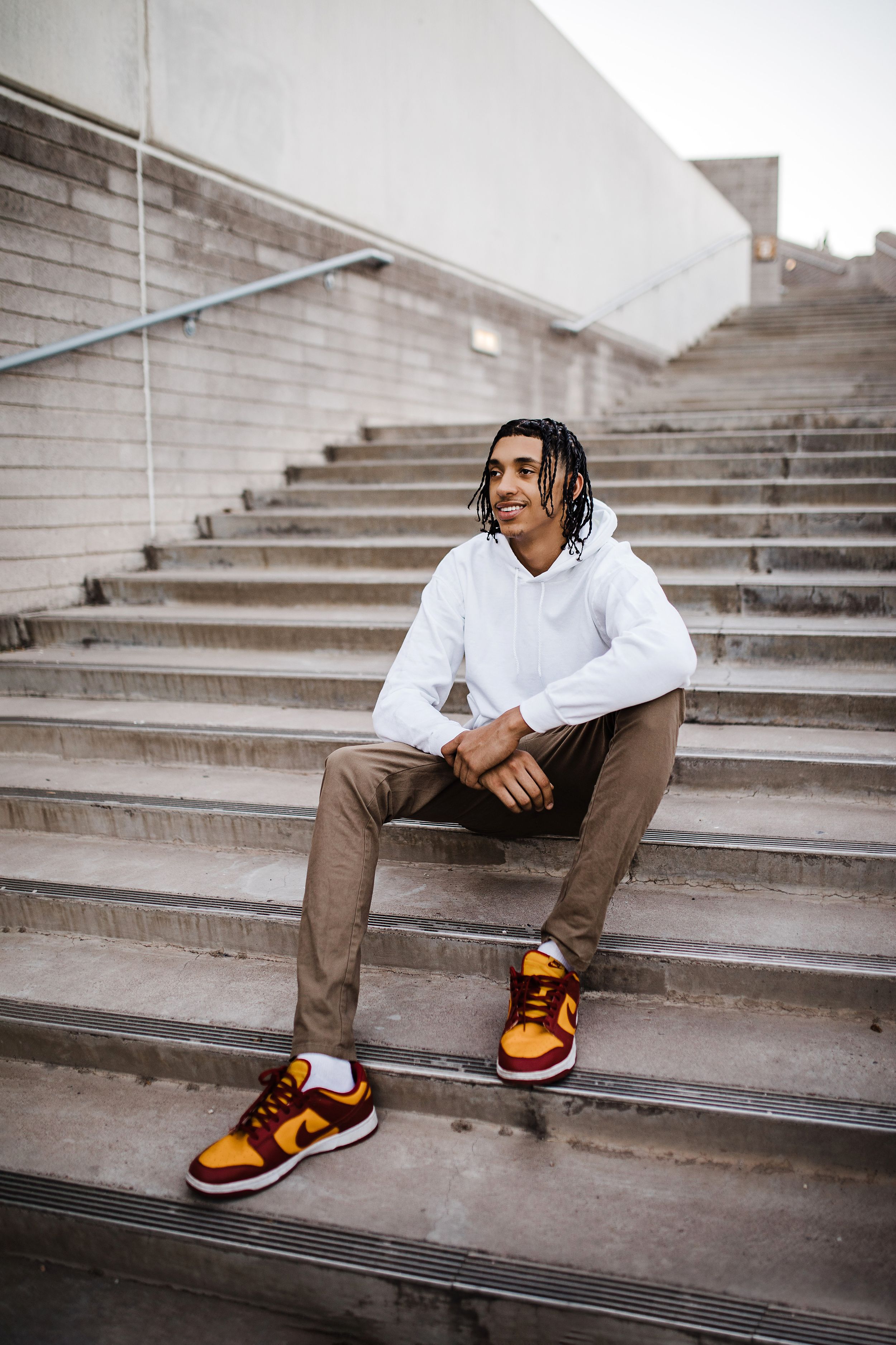 Young man with braids sitting on concrete steps wearing white hoodie and colorful Nike sneakers.