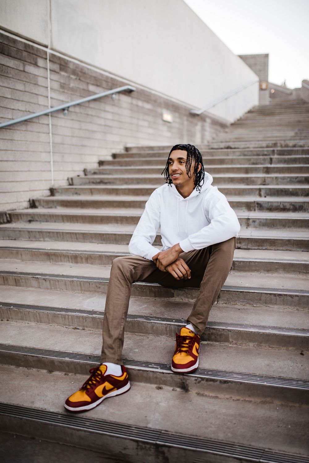 Young man with braids sitting on concrete steps wearing white hoodie and colorful Nike sneakers.
