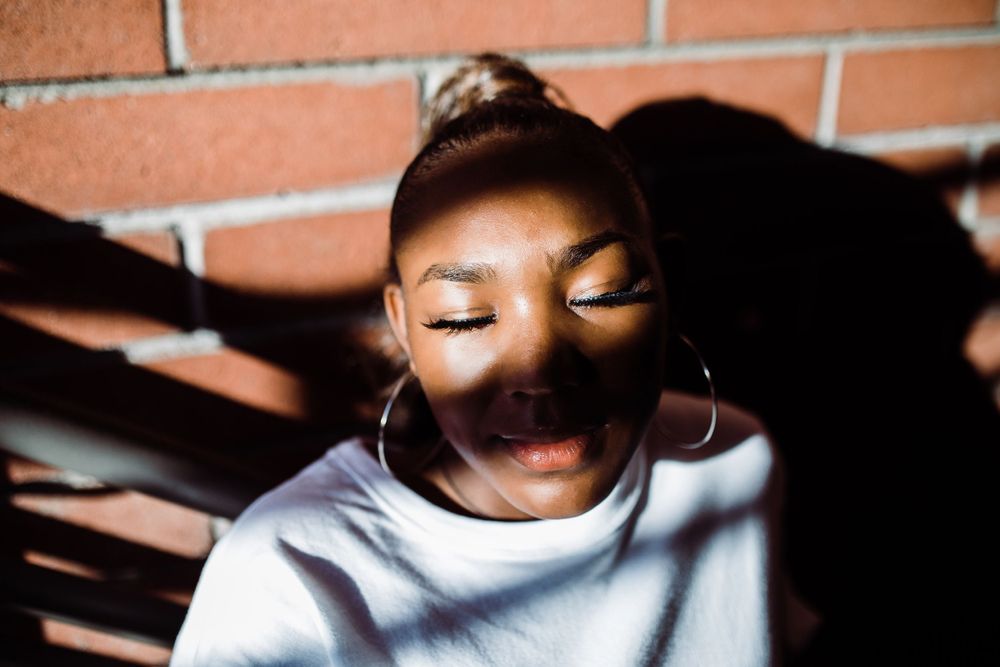 Young Black woman with hoop earrings closes eyes in sunlight against brick wall with dramatic shadows.