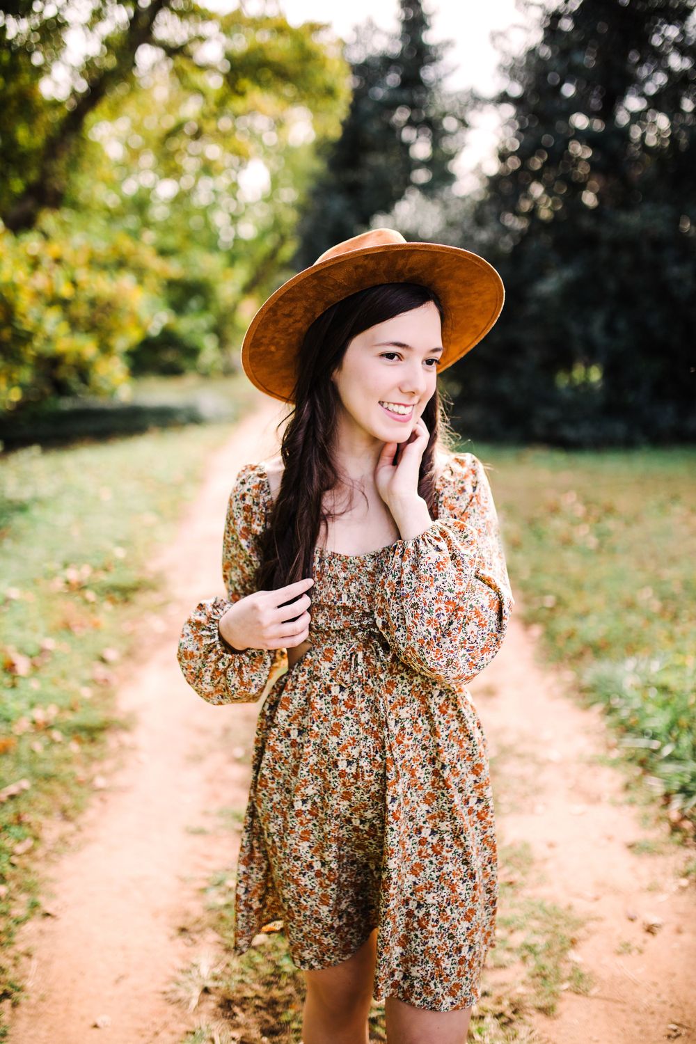 Smiling young woman in floral dress and wide-brim hat on autumn garden path with trees.