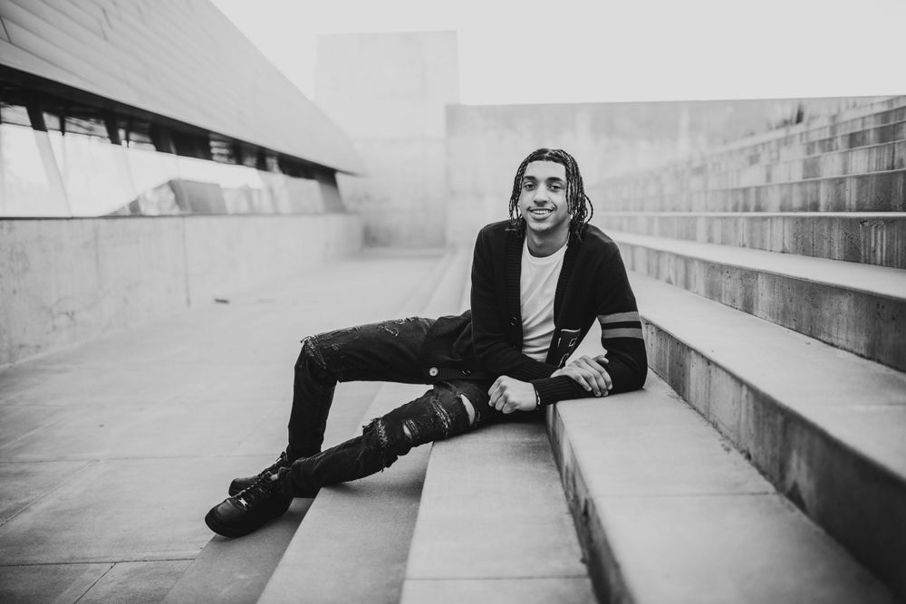 Young man with braids in black cardigan sits relaxed on outdoor steps in black and white photo.