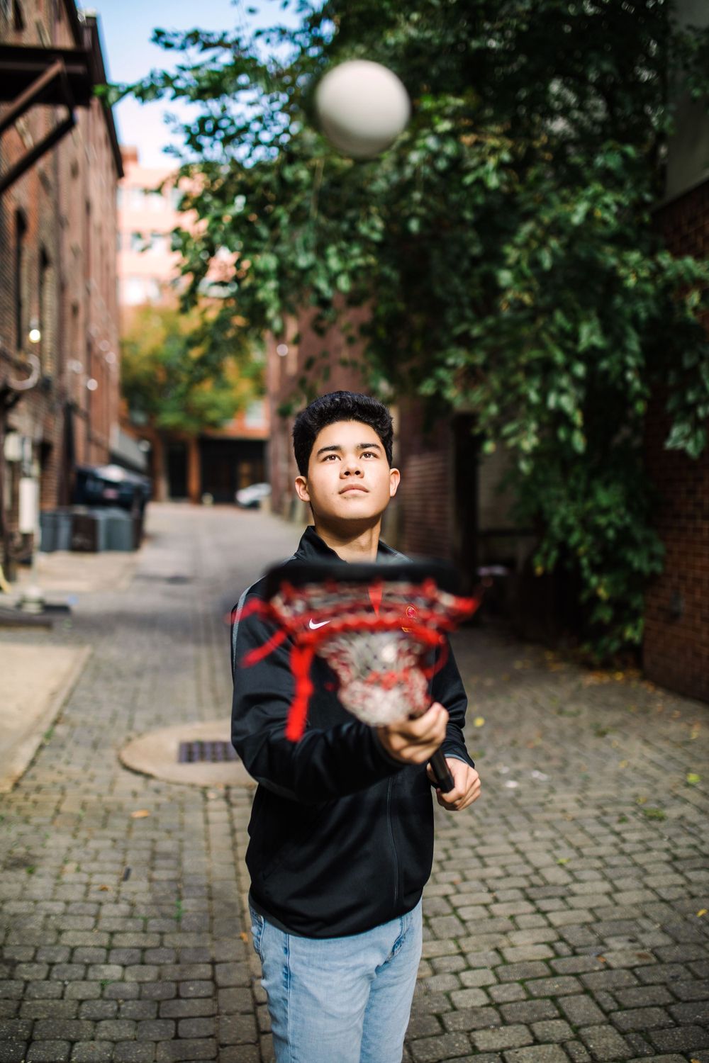 Teen boy tossing lacrosse ball in urban brick alleyway while holding lacrosse stick.