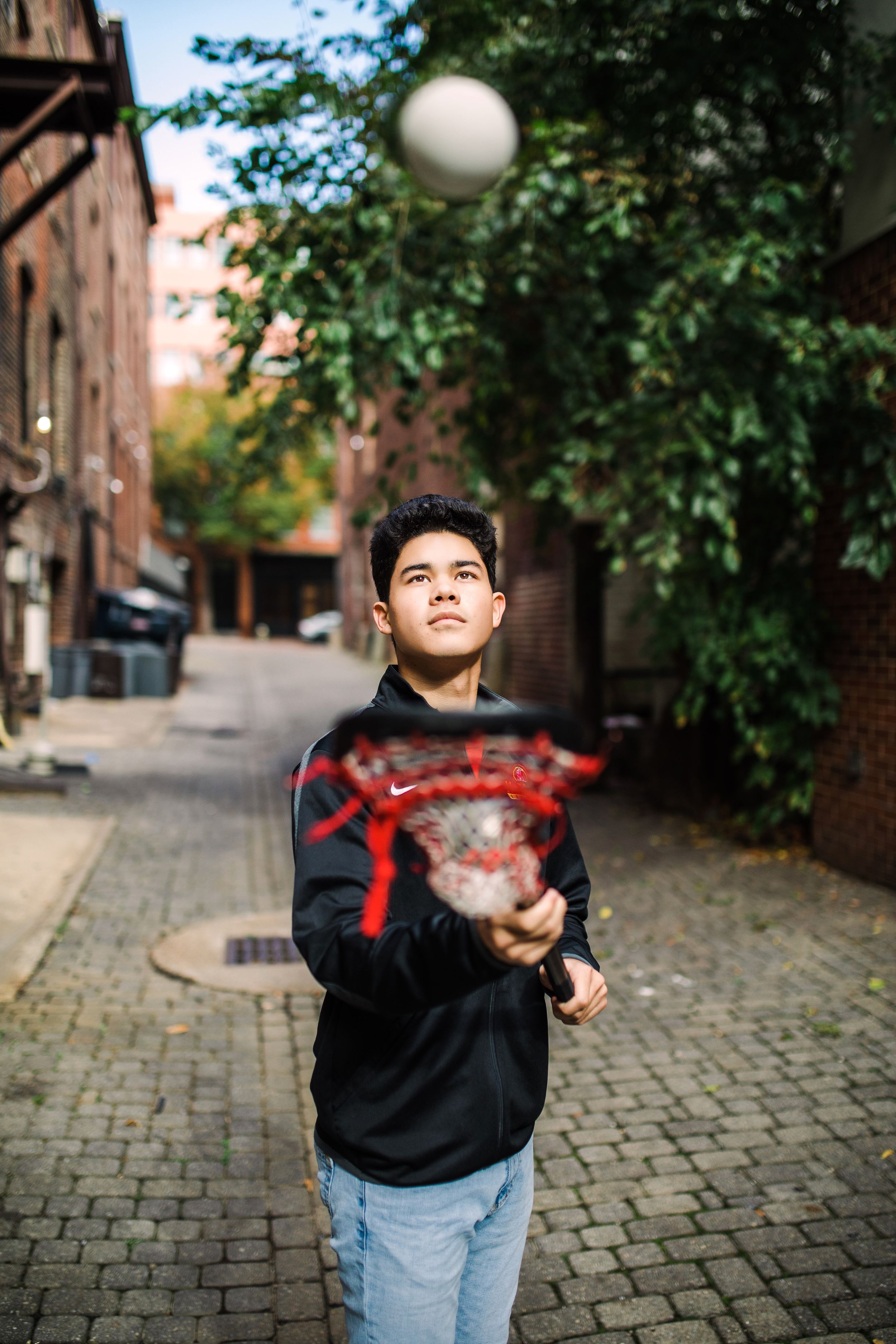 Teen boy tossing lacrosse ball in urban brick alleyway while holding lacrosse stick.