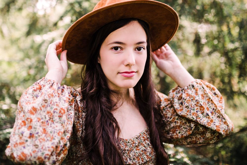 Young brunette woman in floral dress adjusting her wide-brim brown felt hat outdoors in nature.