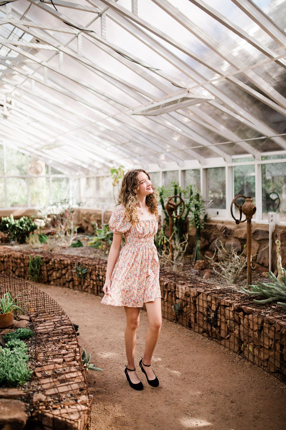 Young woman in floral dress walking through sunlit greenhouse with cacti and stone-walled planters.