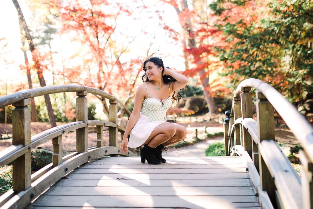 Teen girl in white dress crouching on wooden bridge surrounded by red autumn foliage.