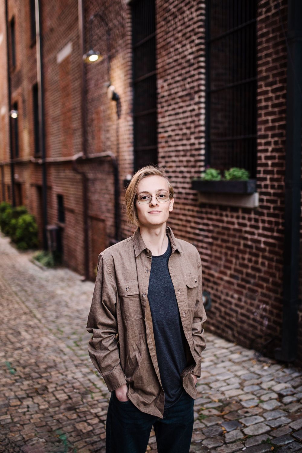 Young man with glasses and brown jacket standing on cobblestone alley beside brick building walls.