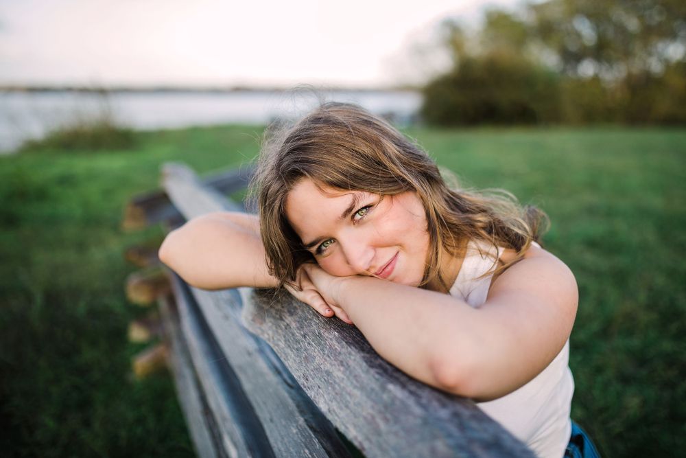 Smiling young woman rests chin on arms leaning on wooden fence near water at sunset.