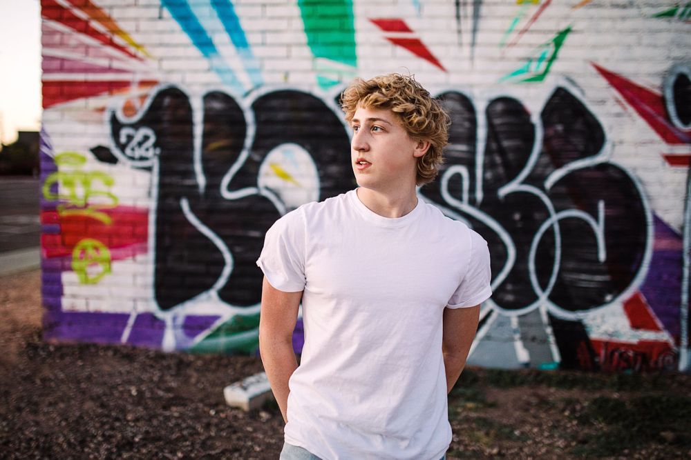 Teen boy in white t-shirt standing before colorful graffiti wall outdoors.