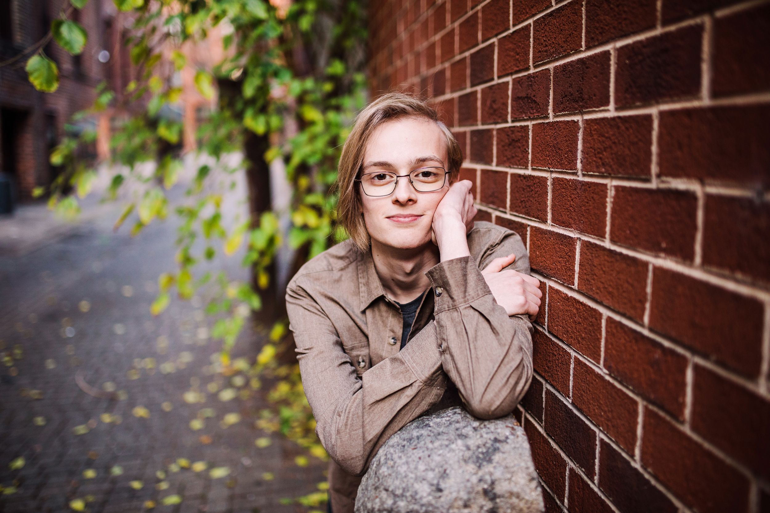 Young person with glasses resting against red brick wall in a green ivy-lined urban alleyway.