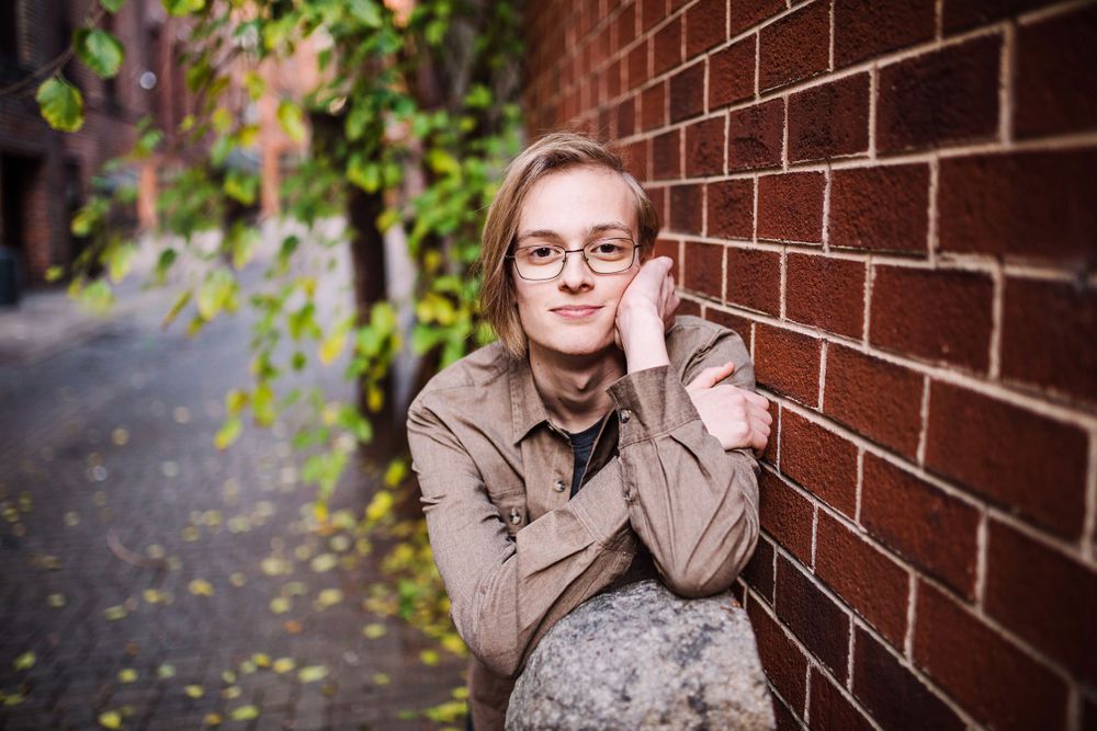 Young person with glasses resting against red brick wall in a green ivy-lined urban alleyway.