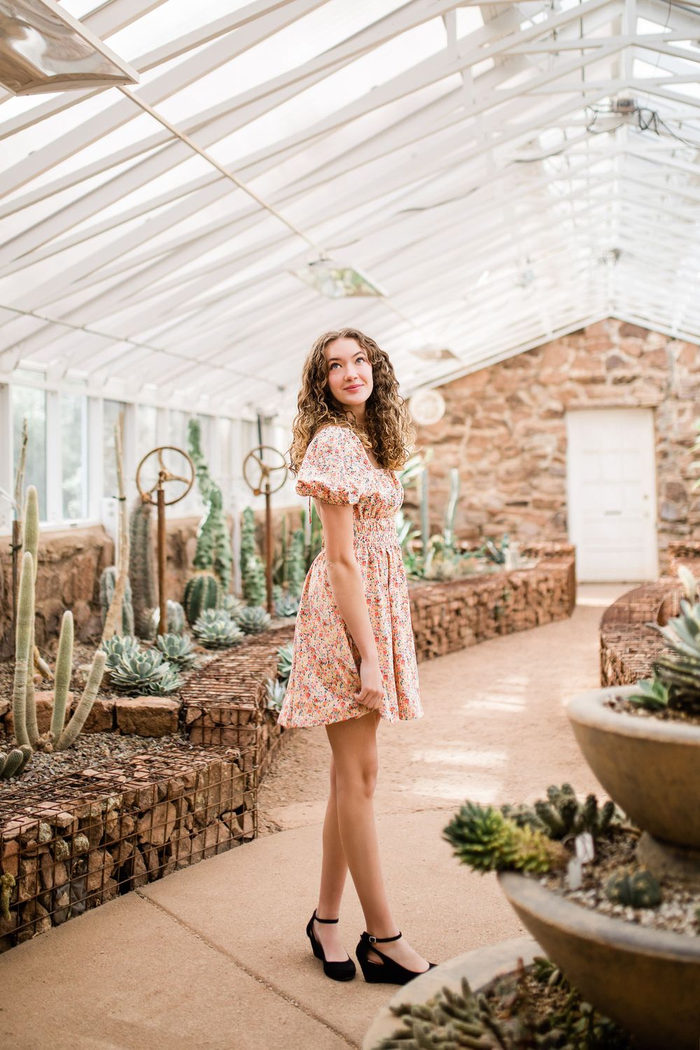 Young woman in floral dress posing inside sunlit greenhouse surrounded by cacti and succulents.