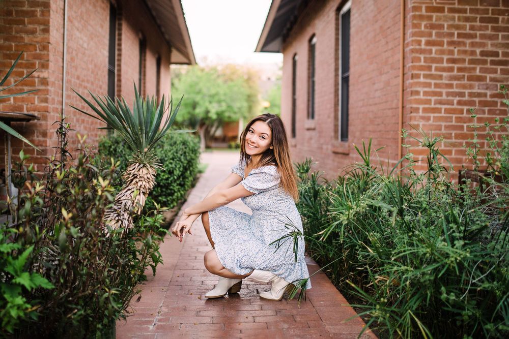 Young woman crouching gracefully on a brick pathway surrounded by lush garden plants.
