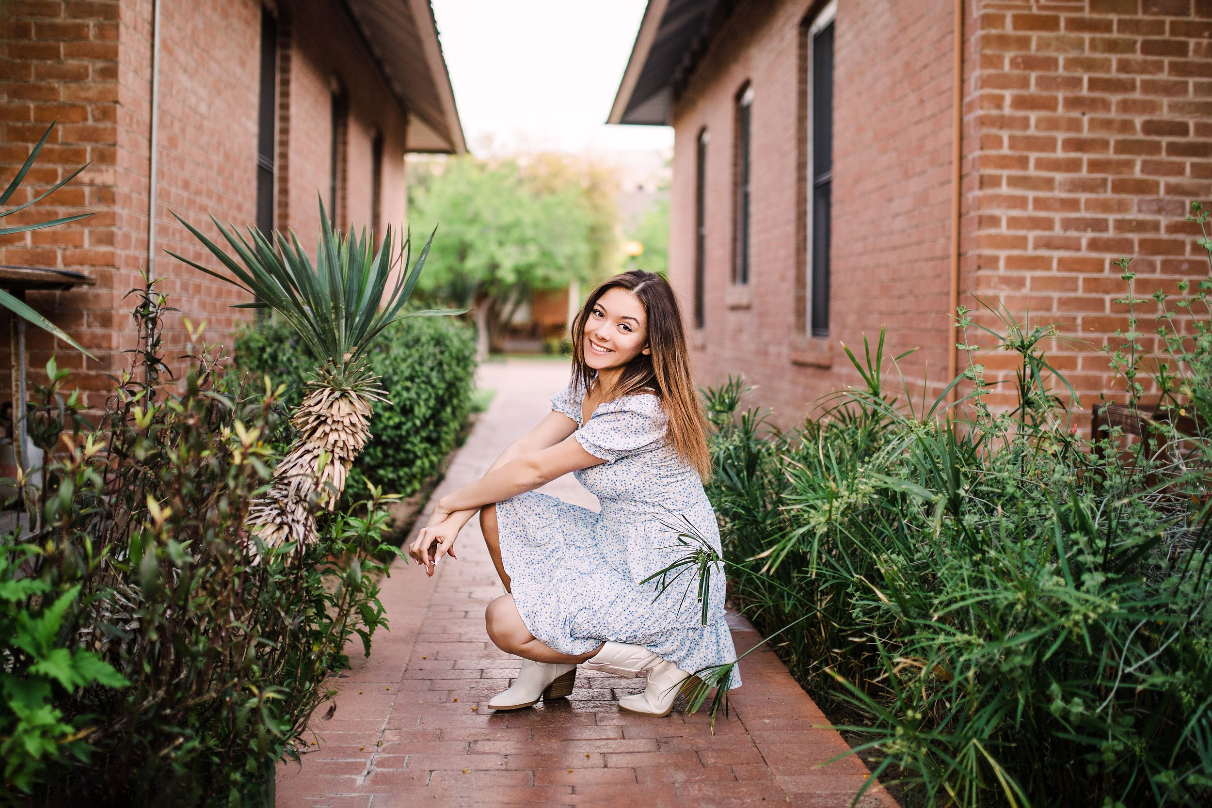 Young woman crouching gracefully on a brick pathway surrounded by lush garden plants.