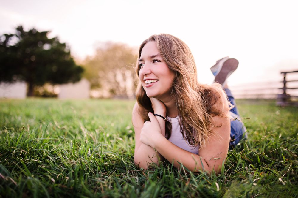 Happy young woman lying on lush green grass smiling in warm golden hour sunlight.