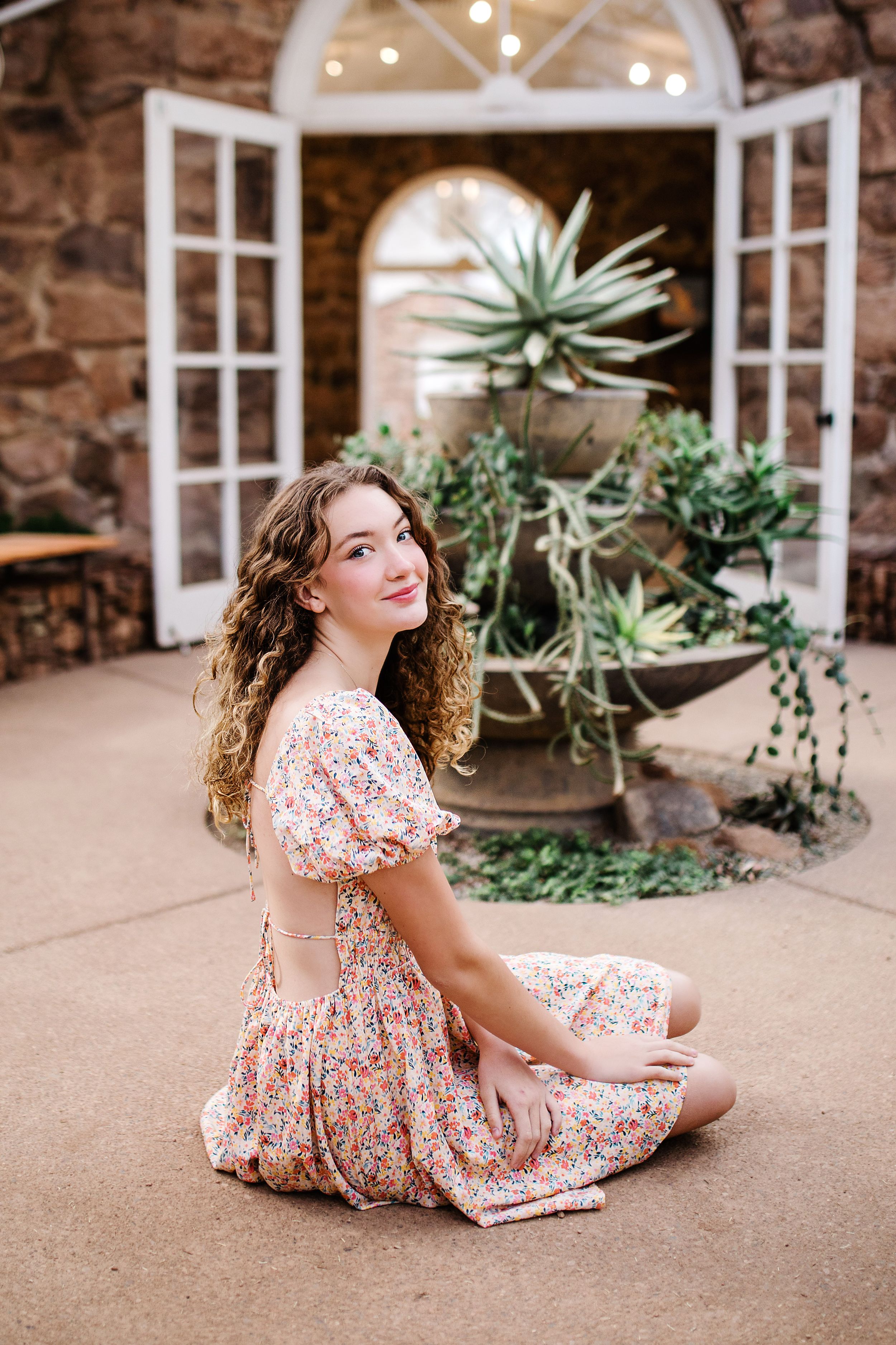 Smiling young woman in floral dress sitting cross-legged in front of greenhouse with succulents.