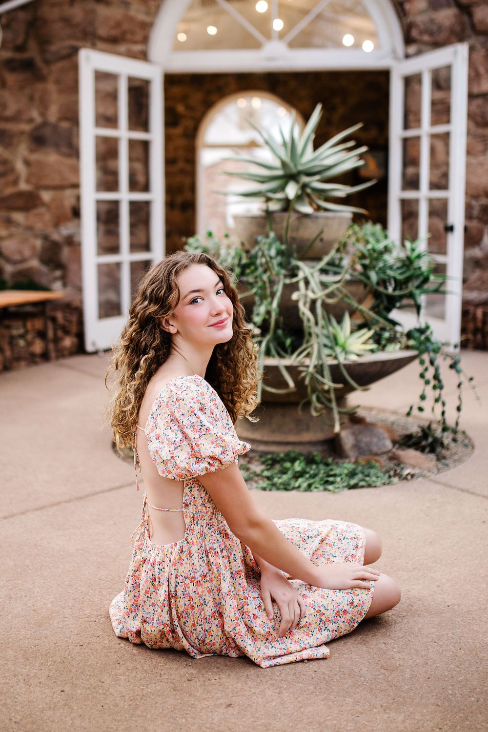 Smiling young woman in floral dress sitting cross-legged in front of greenhouse with succulents.