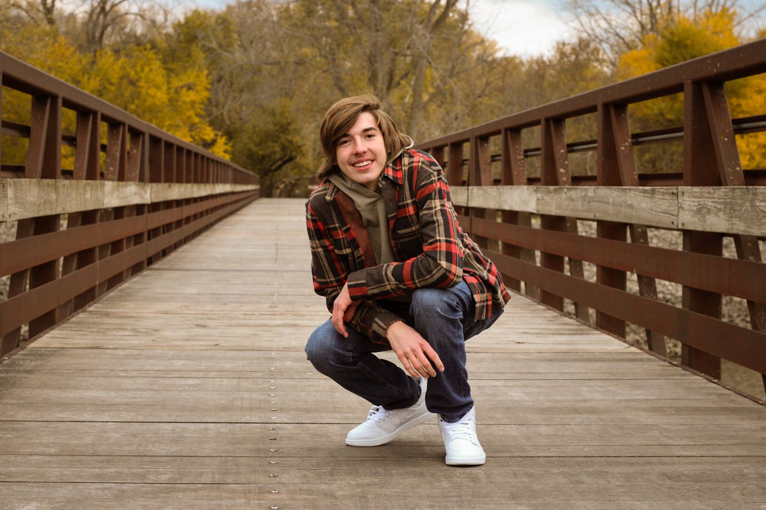 Person in a plaid shirt crouches on a wooden bridge surrounded by autumn foliage.