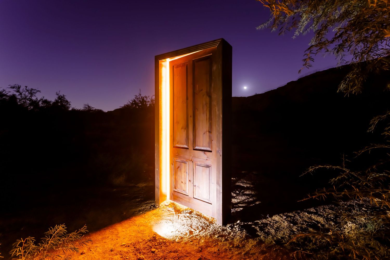 Wooden door emitting warm light stands mysteriously in nighttime desert landscape.