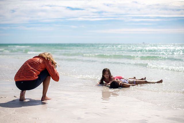 Woman crouching on sandy beach watching young girl lying in shallow ocean waves on a sunny day.