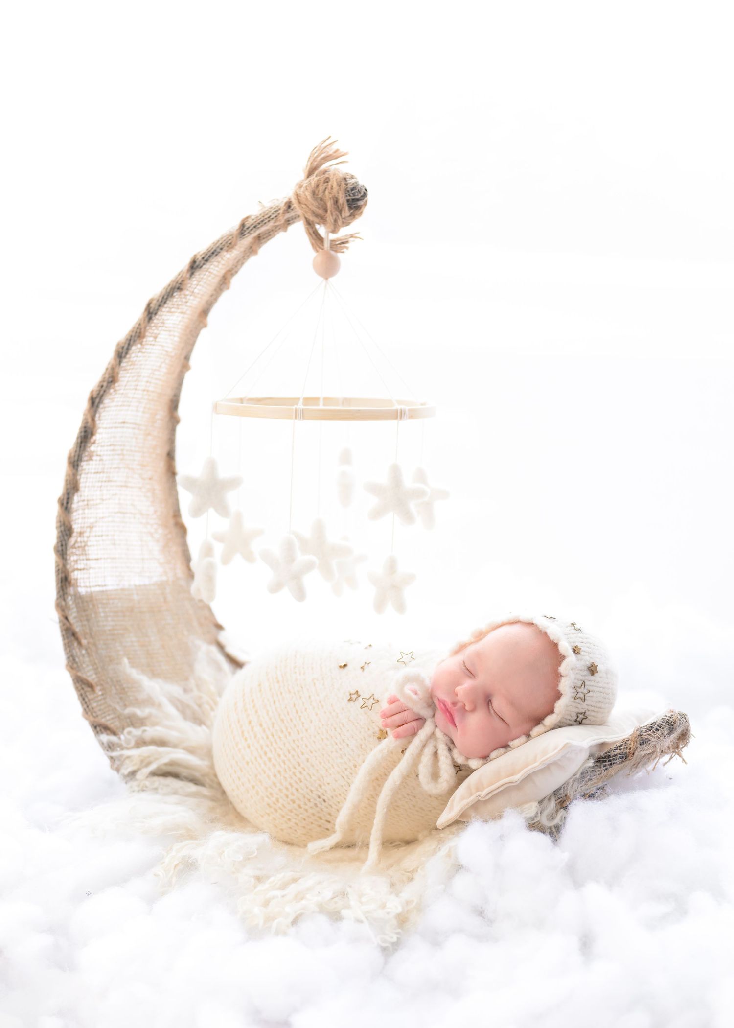 Sleeping newborn wrapped in cream knit blanket rests on decorative crescent moon prop with soft cloud-like backdrop.