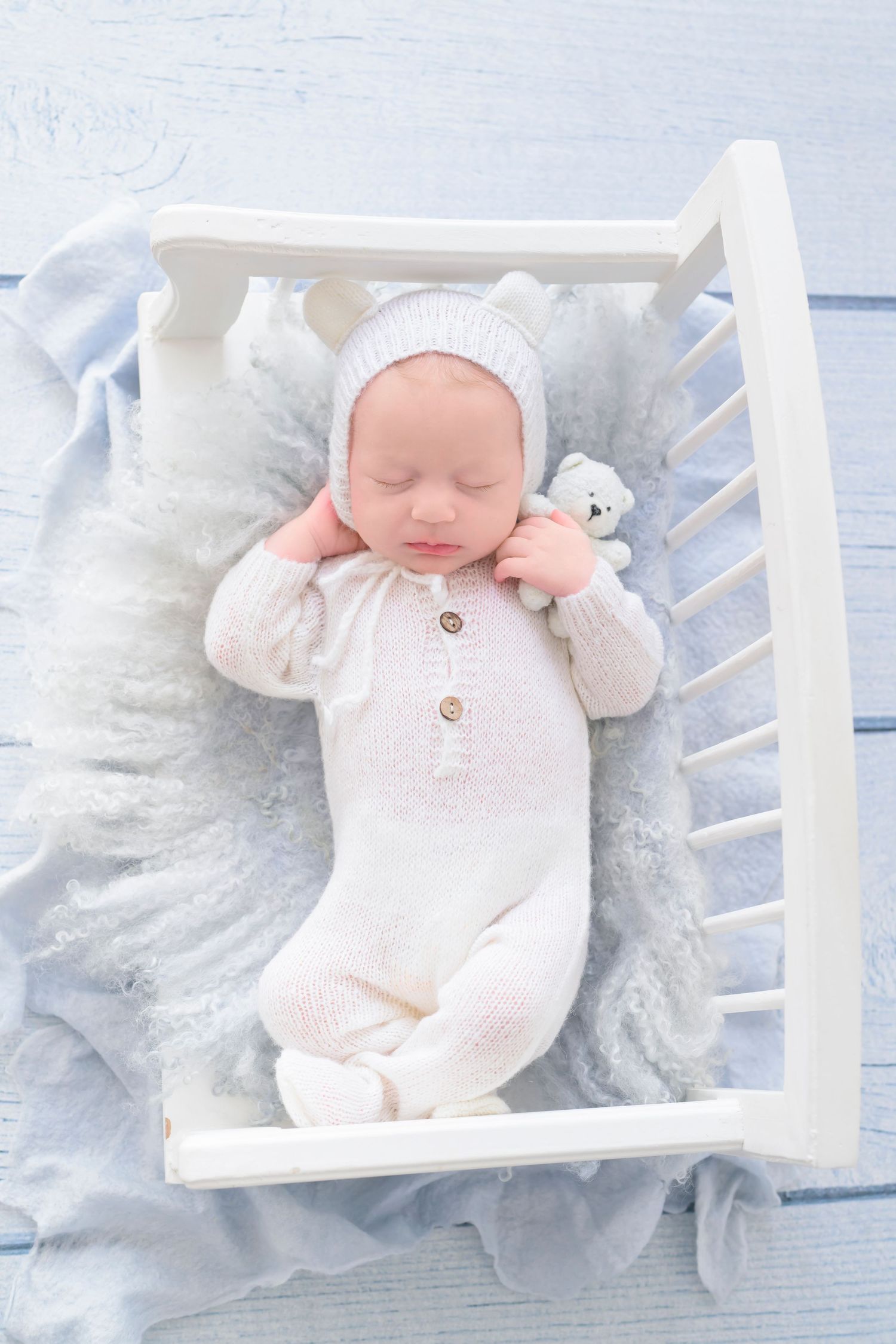 A sleeping baby in a white knit outfit lies peacefully in a white crib with soft blue bedding.