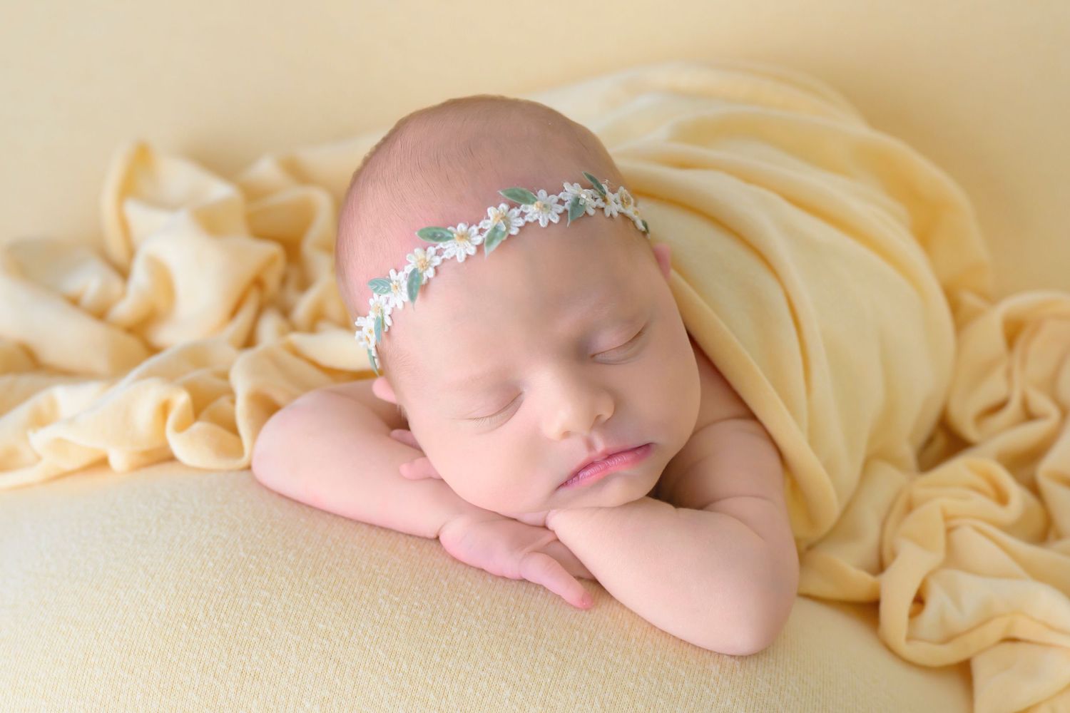 Peaceful sleeping newborn wrapped in soft yellow fabric wearing delicate crystal headband rests on baby yellow backdrop.