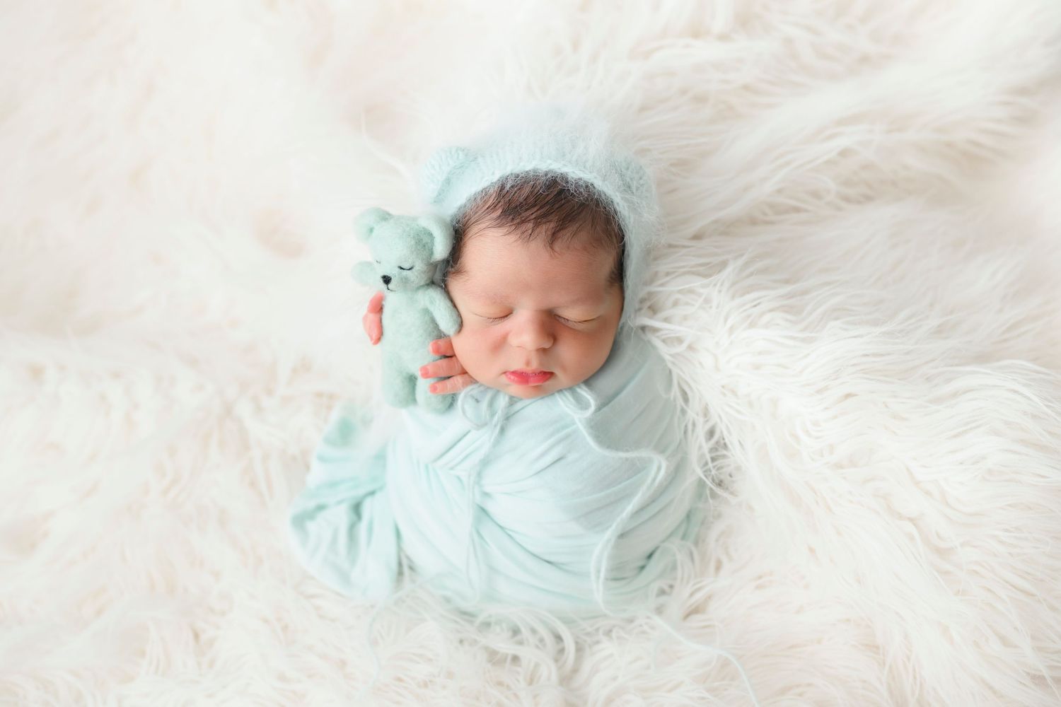 A sleeping newborn wrapped in mint green swaddle with matching plush toy rests on soft white fur blanket.