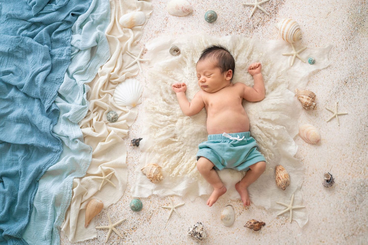 Newborn baby sleeping on sandy beach setup surrounded by seashells and starfish wearing teal shorts.