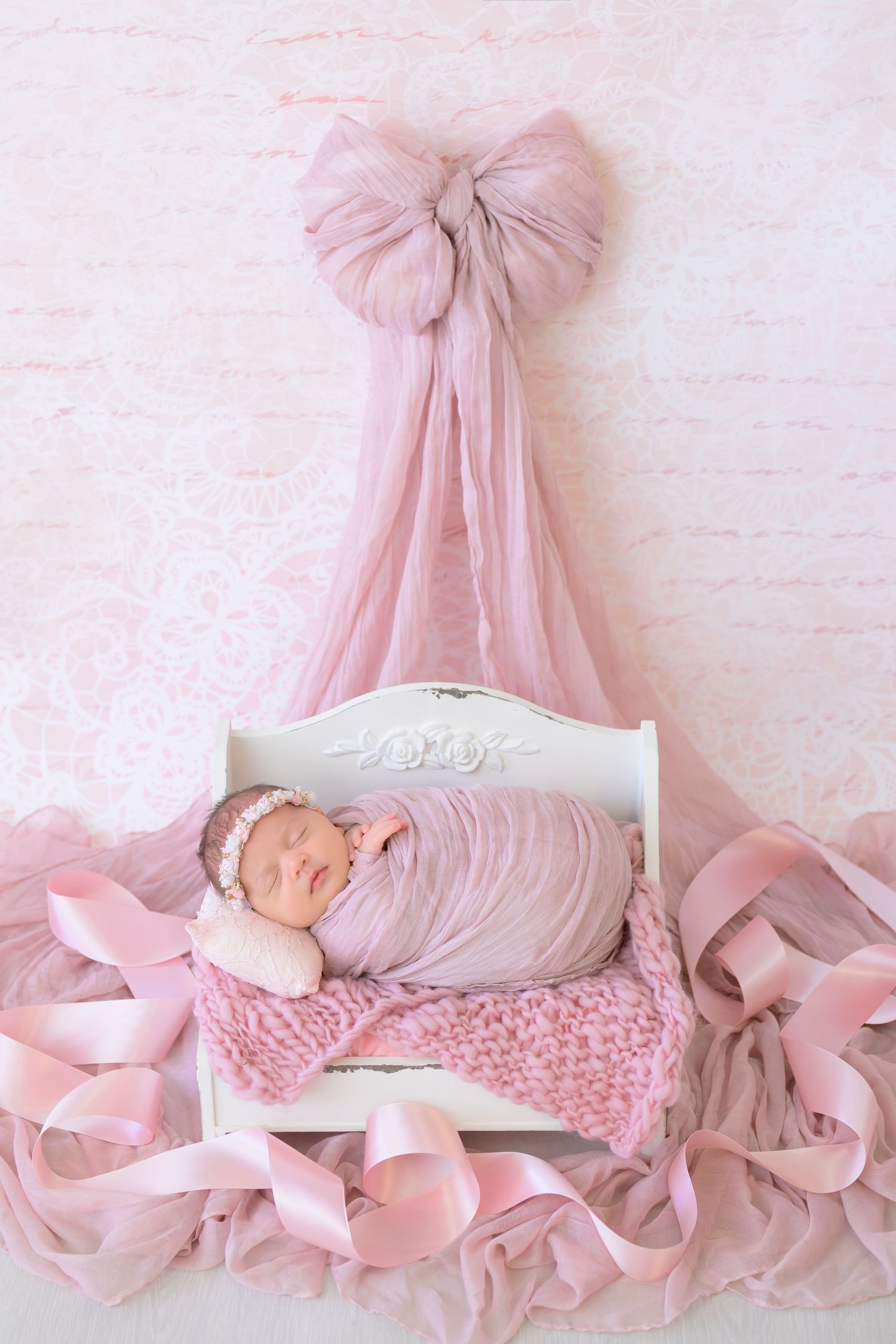Newborn sleeping in white vintage bed decorated with soft pink bows and tulle backdrop in romantic nursery setting.