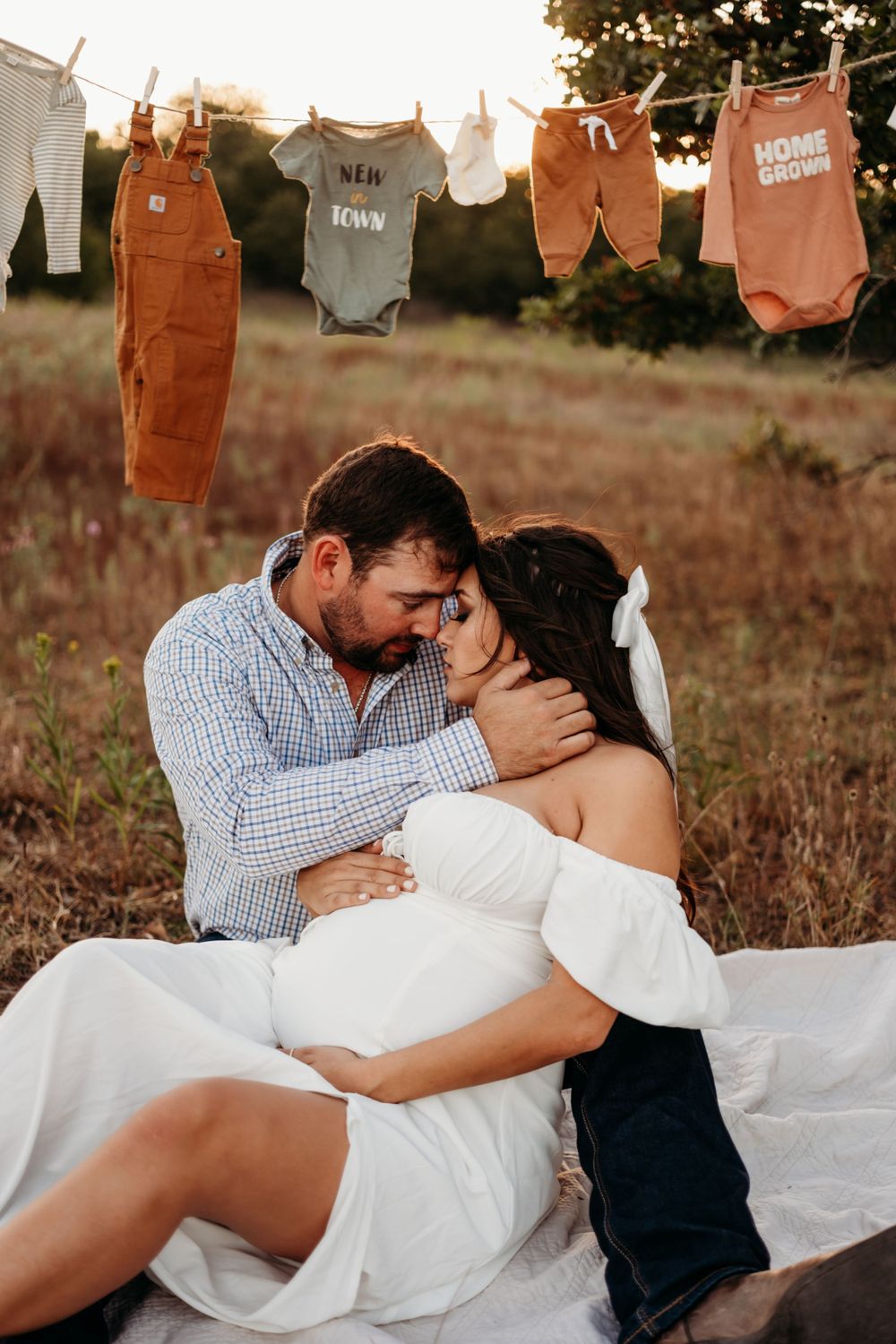 Baby clothes on clothesline with couple embracing below during sunset photoshoot.