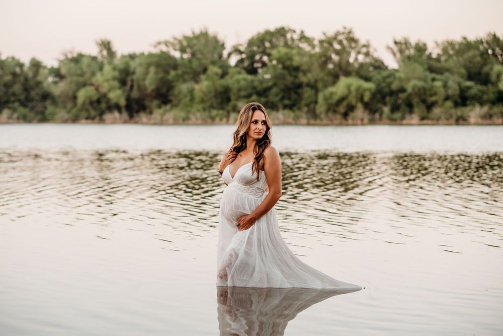 An ethereal maternity photo shows a white dress flowing in water during sunset by a lake.