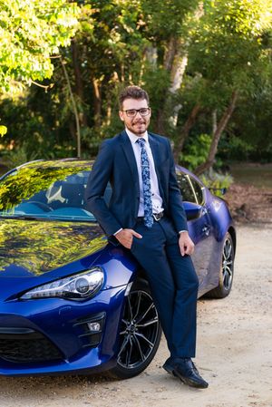 Professional portrait shoot featuring a blue sports car and person in navy suit against natural outdoor backdrop.
