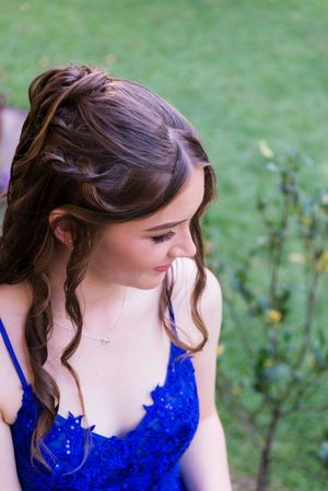 A woman in a royal blue lace dress stands outdoors with an elegant updo hairstyle and curled tendrils.