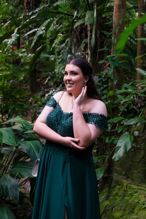 Woman in emerald green formal dress poses among lush tropical foliage.