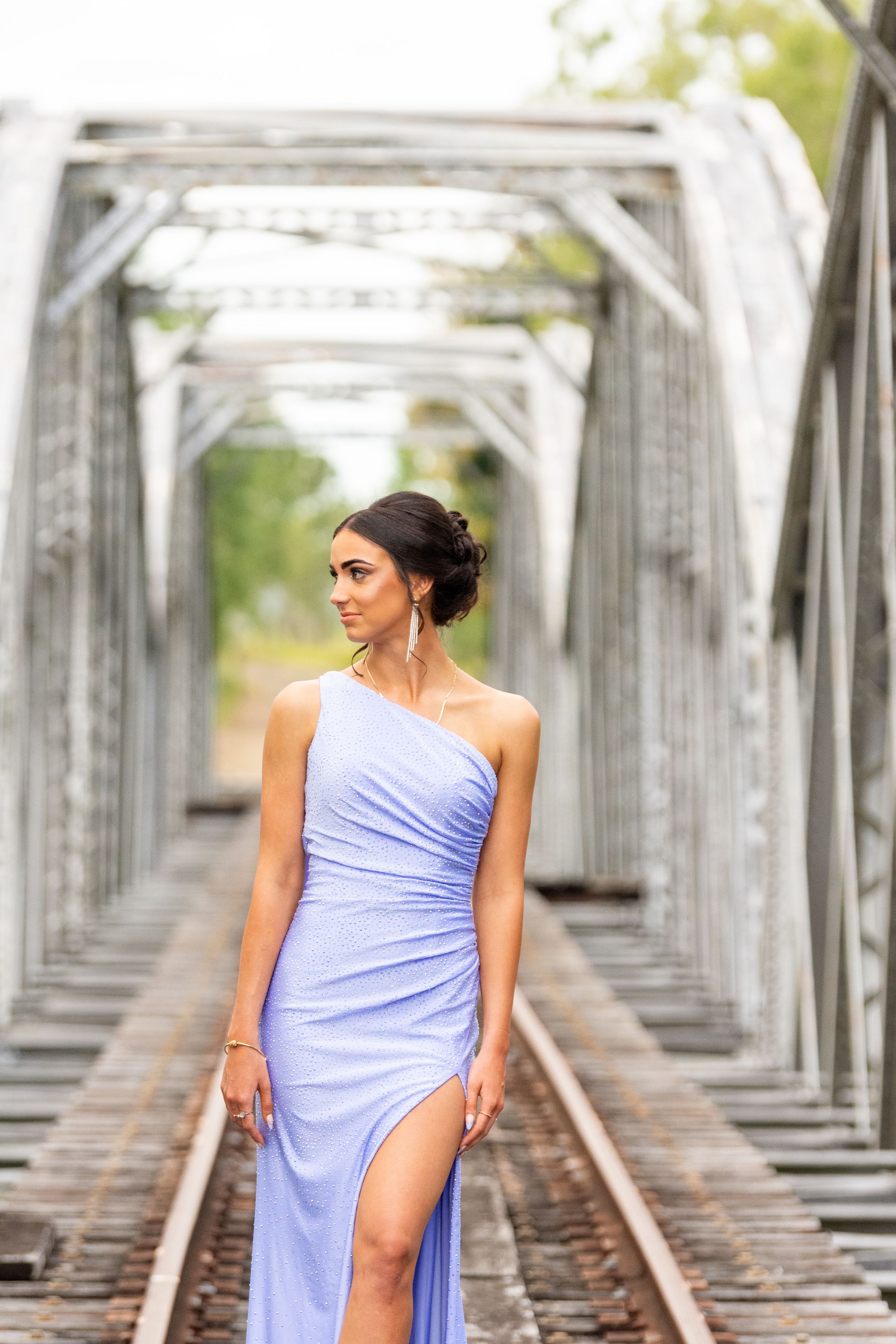 Elegant model in flowing lilac gown poses on vintage railway bridge.