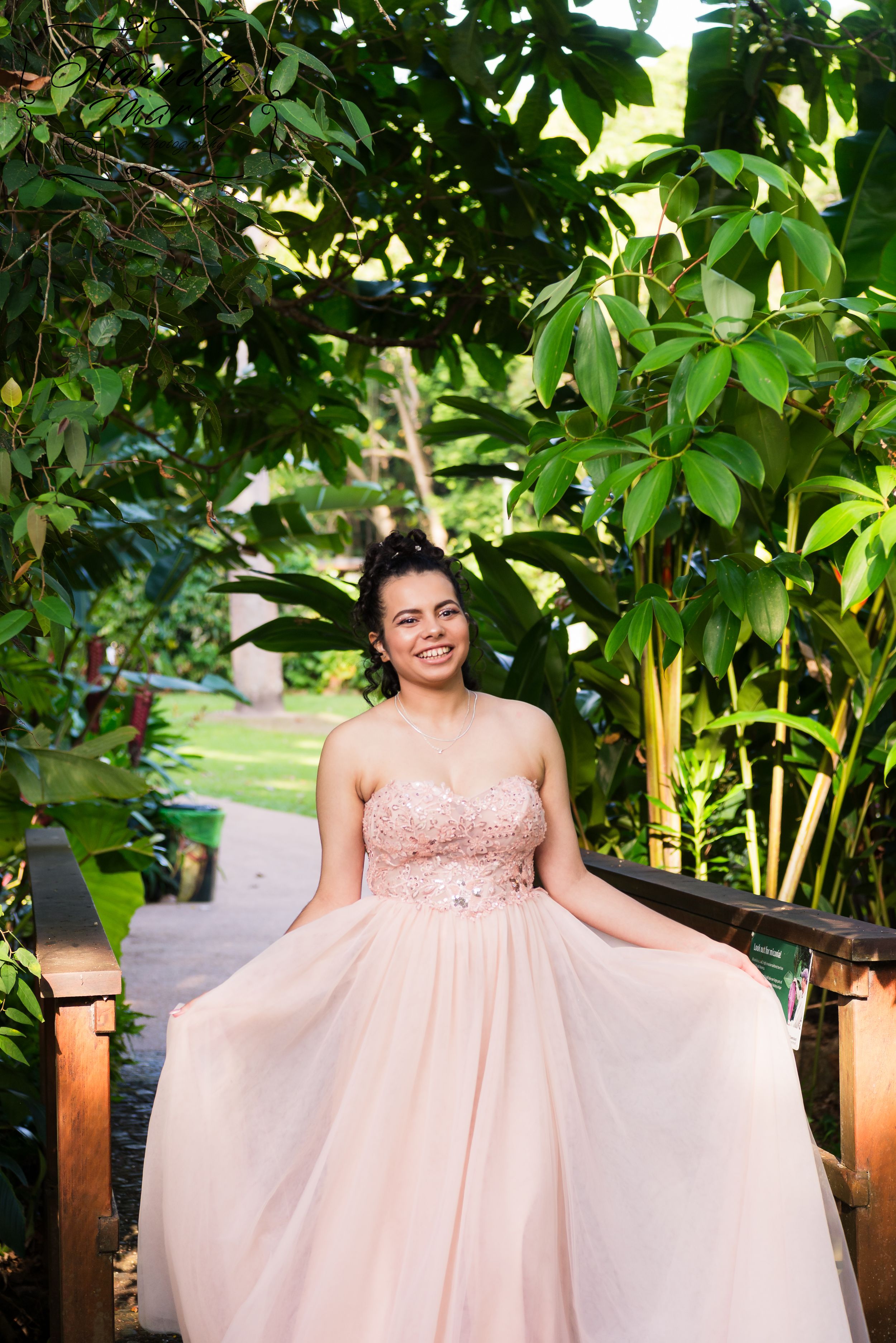 A smiling person in a blush pink formal gown poses in front of lush tropical foliage and bamboo.