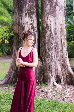 Woman in burgundy dress poses against large tree trunks in garden setting.