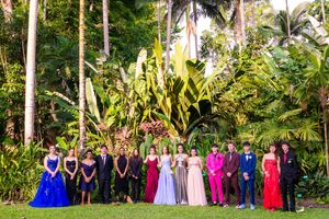 Large formal group photo with people in colorful formal attire against tropical garden backdrop.