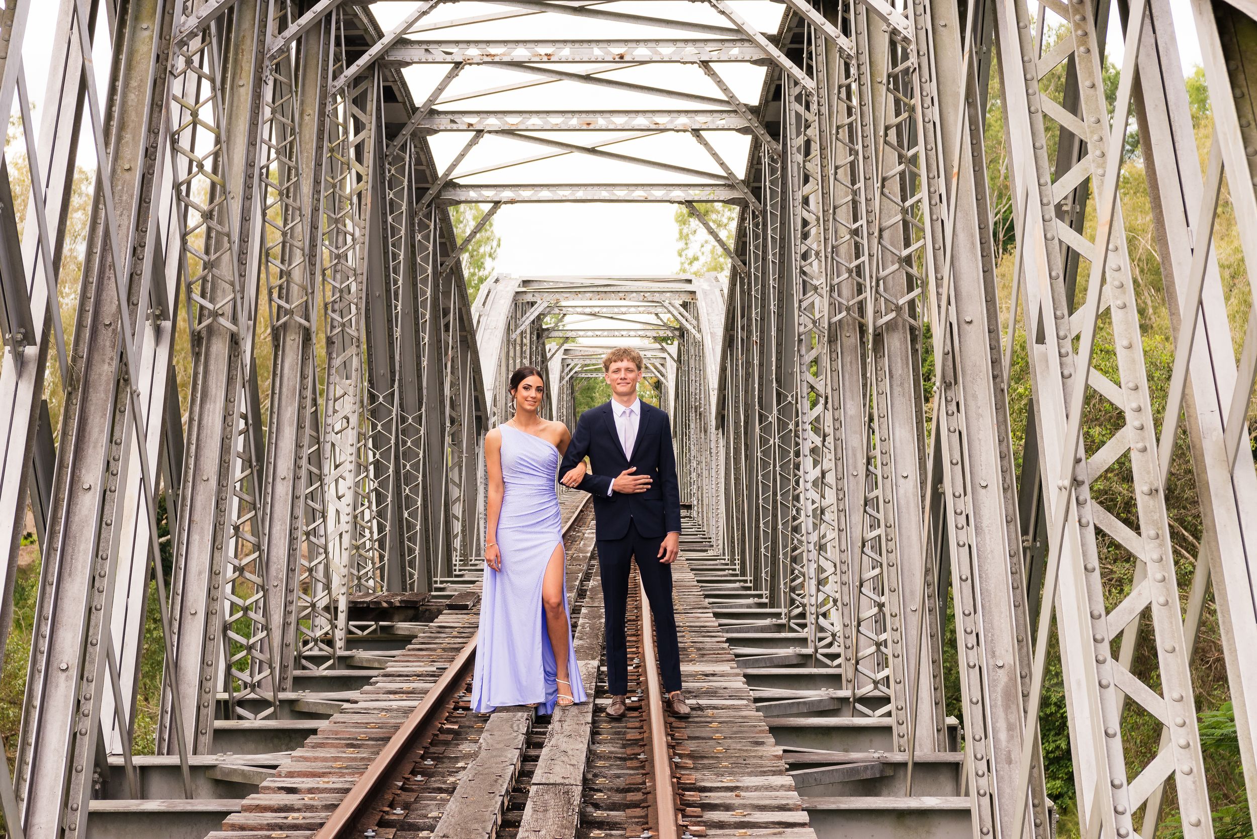 Long view of a couple walking on rustic railroad bridge framed by metal trusses.
