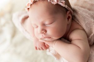 A sleeping newborn baby wearing a pink headband rests peacefully on soft white bedding.