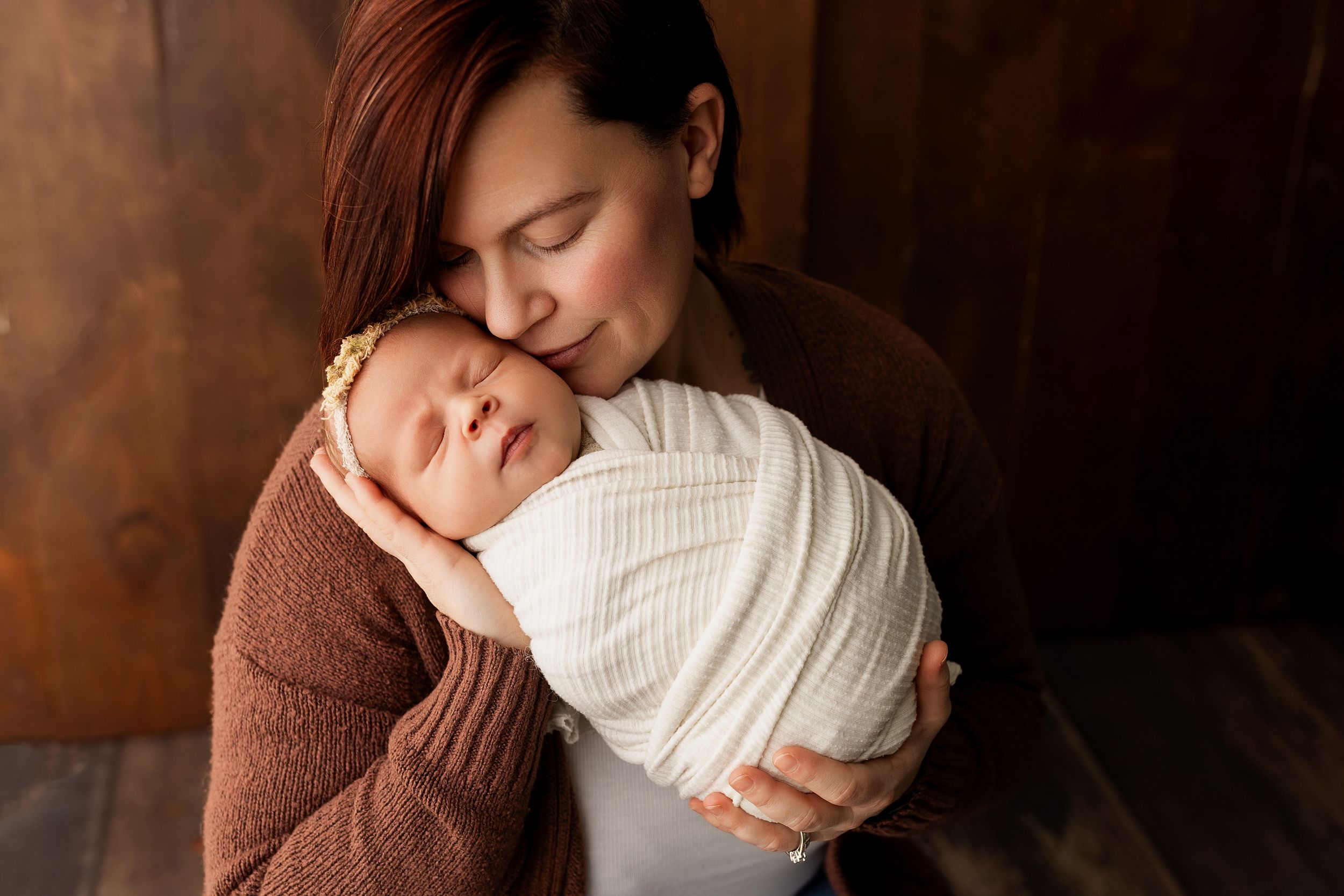 A newborn baby swaddled in white fabric is held tenderly against a brown sweater in a warm-toned portrait.