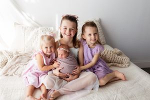 Children wearing pastel colored outfits sit together on a white bed with plush pillows and blankets.