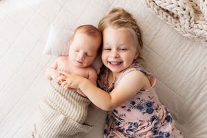 A newborn baby and toddler sibling lie together on white bedding while sharing sweet moments and smiles.