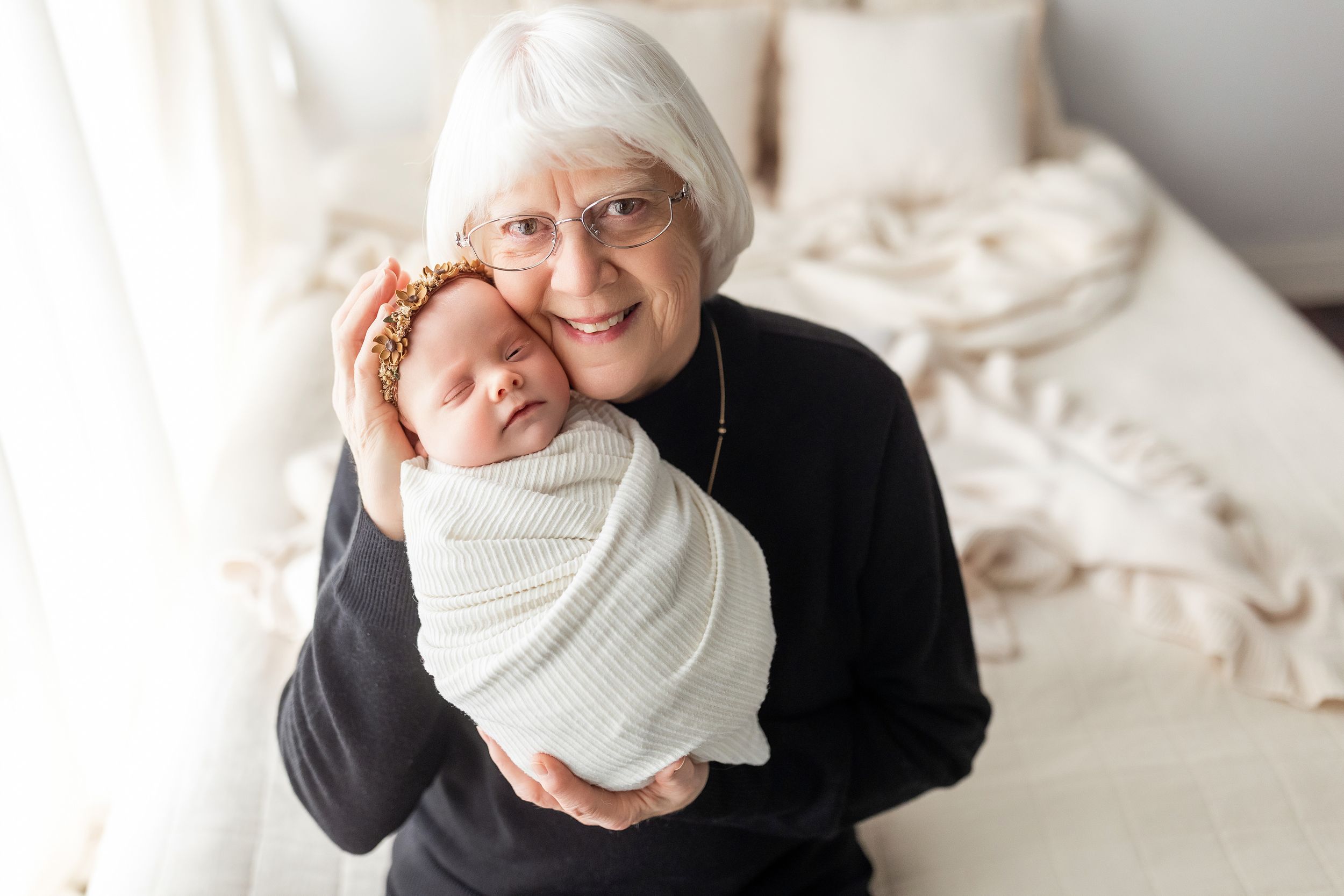 A tender moment shared between a woman in black and a swaddled newborn baby on a white bed.