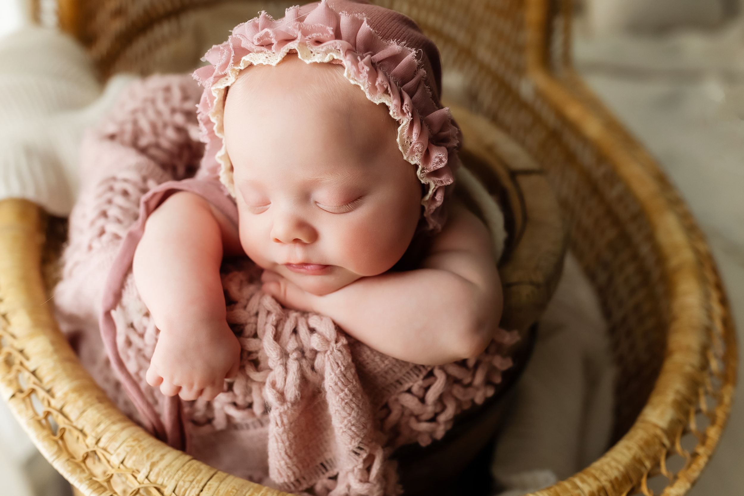 Sleeping newborn wearing pink lace bonnet rests peacefully in vintage woven basket with soft lighting.