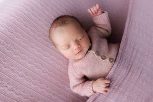 Sleeping baby in pink knit sweater rests on textured mauve bedding.