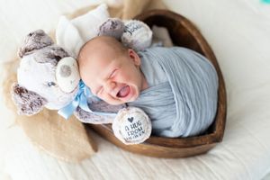 Newborn wrapped in blue swaddle laughs while lying in wooden heart bowl with plush toys.
