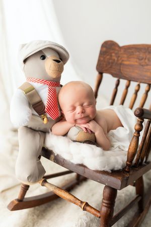 Newborn rests on wooden rocking chair next to decorative teddy bear wearing suspenders and tie.